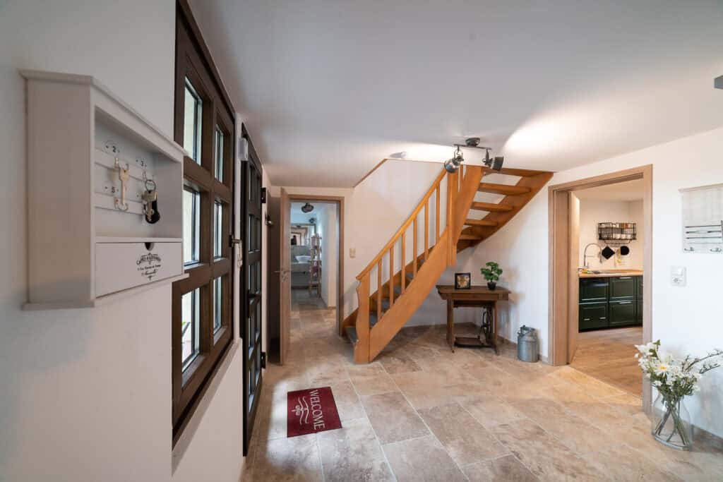 Heller Eingangsbereich eines Ferienhauses in Monschau mit gefliestem Boden, Holztreppe, kleinem Tisch mit Deko, roter Fußmatte und Blick in die Küche – perfekt für ein Eifelwochenende oder eine Eifel-Erkundung mit Kindern. Fenster und Schlüsselbrett an der linken Wand.