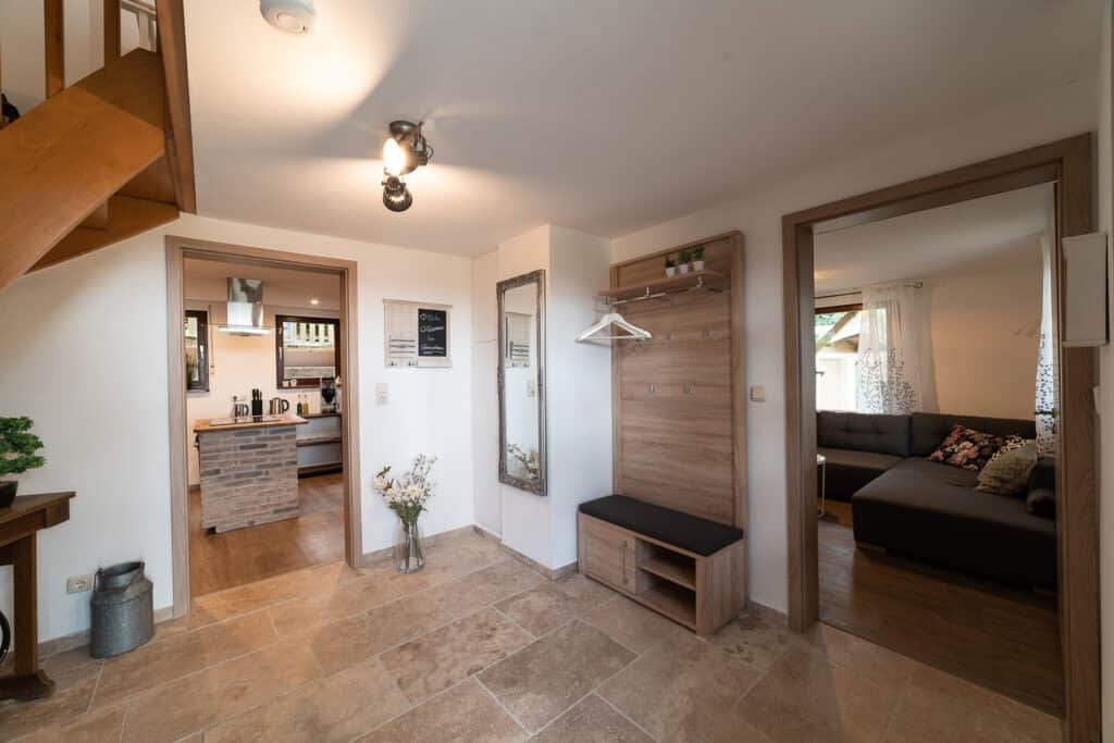 Heller Eingangsbereich mit Fliesenboden, Holztreppe, Spiegel, Sitzbank, Kleiderhaken und Blick in die moderne Küche des Ferienhauses Eifel links und ein gemütliches Wohnzimmer mit grauem Sofa rechts – perfekt für Familien, die die Eifel mit Kindern erkunden.
