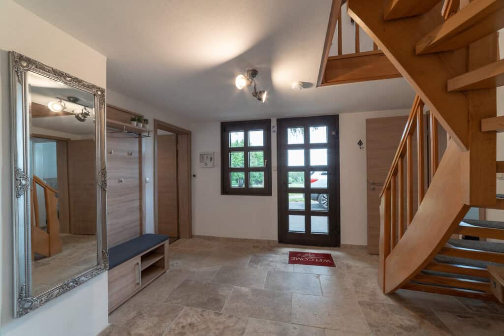 Ein heller Eingangsbereich begrüßt Sie in diesem Ferienhaus Eifel mit Fliesenboden, großem Wandspiegel, Holztreppe, Garderobe mit Sitzbank und Glastür. Eine rote „Willkommen“-Matte begrüßt die Gäste, während das Sonnenlicht durch die Fenster hereinströmt.