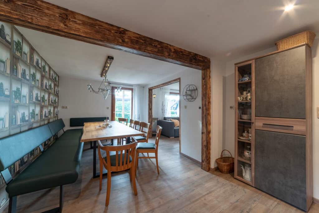Ein gemütlicher Essbereich im Monschau Ferienhaus mit Holztisch, gemischten Stühlen und einer grünen Bank. Kunstwerke und Regale mit Gläsern schmücken die Wände. Der Raum öffnet sich zu einem hellen Wohnzimmer – ideal für einen Familienurlaub in der Eifel.