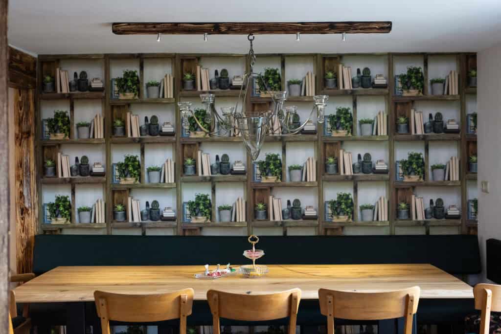 Ein hölzerner Esstisch mit sechs Stühlen steht unter einem Kronleuchter vor einer Wand, die mit Bücherregalen, Topfpflanzen und kleinen Dekoartikeln dekoriert ist – perfekt für einen Familienurlaub in der Eifel oder ein gemütliches Eifelwochenende. Auf dem Tisch steht ein mehrstöckiges Tablett mit Süßigkeiten.