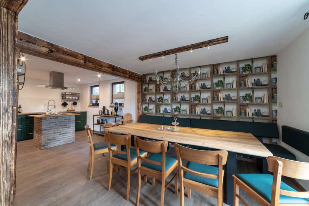 Ein geräumiges Esszimmer mit einem langen Holztisch, umgeben von Stühlen, ideal für einen Familienurlaub in der Eifel. Im Hintergrund schaffen eine moderne Küche und Regale voller Bücher und Pflanzen eine gemütliche Ferienhaus-Atmosphäre in der Eifel, gekrönt von einem rustikalen Kronleuchter.
