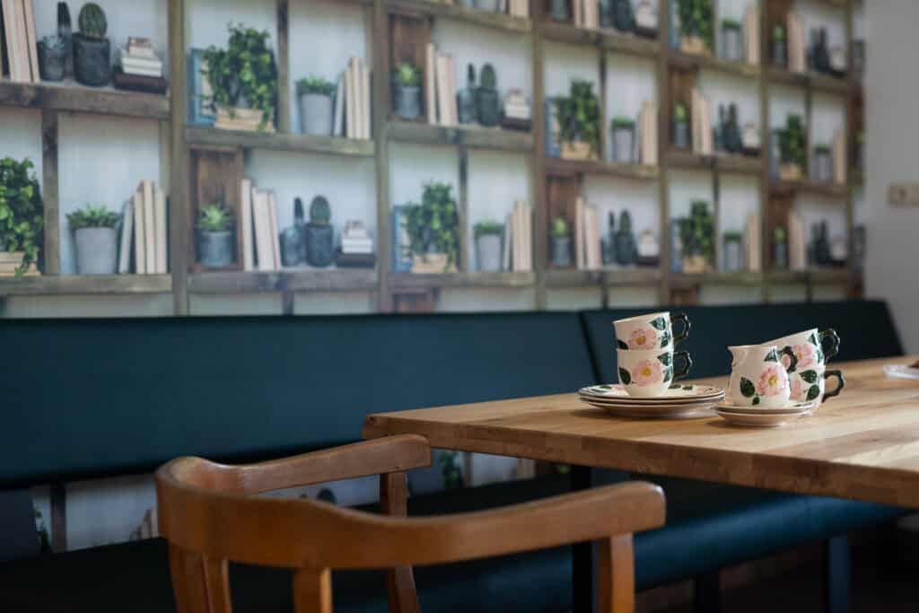Eine gemütliche Café-Ecke mit Holztisch, Stühlen und geblümten Teetassen erwartet Sie – perfekt zum Entspannen nach einem Familienurlaub in der Eifel. Die Tapete zeigt Regale mit Büchern und Topfpflanzen und versprüht den Charme eines echten Ferienhauses in der Eifel.