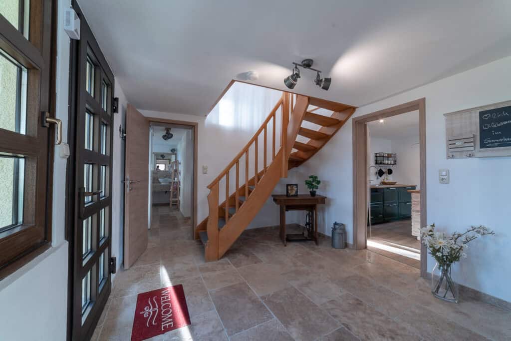 Heller Eingangsbereich mit Steinfliesenboden, Holztreppe und Fußmatte an der Tür – perfekt für ein Monschau Ferienhaus. Ein kleiner Tisch und Dekoartikel stehen unter der Treppe. Zwei offene Türen führen zu weiteren Räumen, darunter eine Küche.