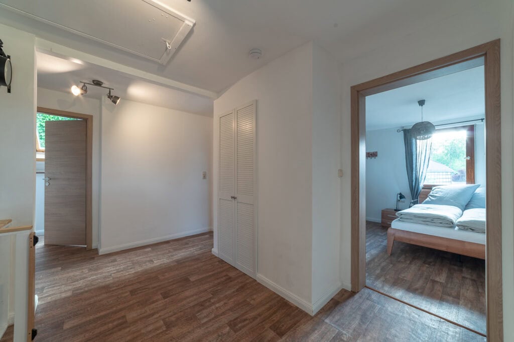 Ein heller Flur mit Holzboden, weißen Wänden und einem Schrank führt zu einem gemütlichen Schlafzimmer mit einem Doppelbett – perfekt für Familien, die die Eifel mit Kindern genießen – in einem Ferienhaus Eifel mit großen Fenstern und luftigen, durchsichtigen Vorhängen.