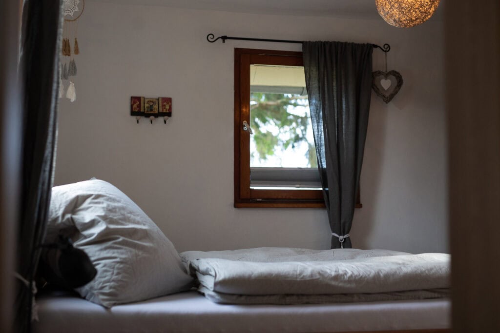 Ein kleines, gemütliches Schlafzimmer mit einem Einzelbett, grauer Bettwäsche und einem Kissen befindet sich unter einem Fenster mit dunklen Vorhängen. Perfekt für Familien, die die Eifel mit Kindern erkunden oder in einem Ferienhaus mit Sauna in der Eifel entspannen. Sanftes Licht und Wanddekoration sorgen für eine heimelige Atmosphäre.