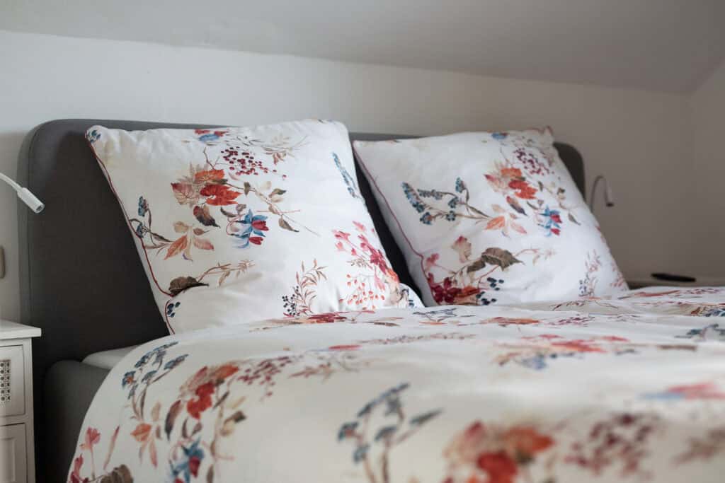 Ein ordentlich gemachtes Bett mit weißen Laken und geblümten Kissenbezügen steht vor einem grauen Kopfteil in einem gemütlichen, sanft beleuchteten Schlafzimmer – perfekt zum Entspannen nach einem Tag in der Eifel oder zum Abschalten im Ferienhaus Sauna Eifel.