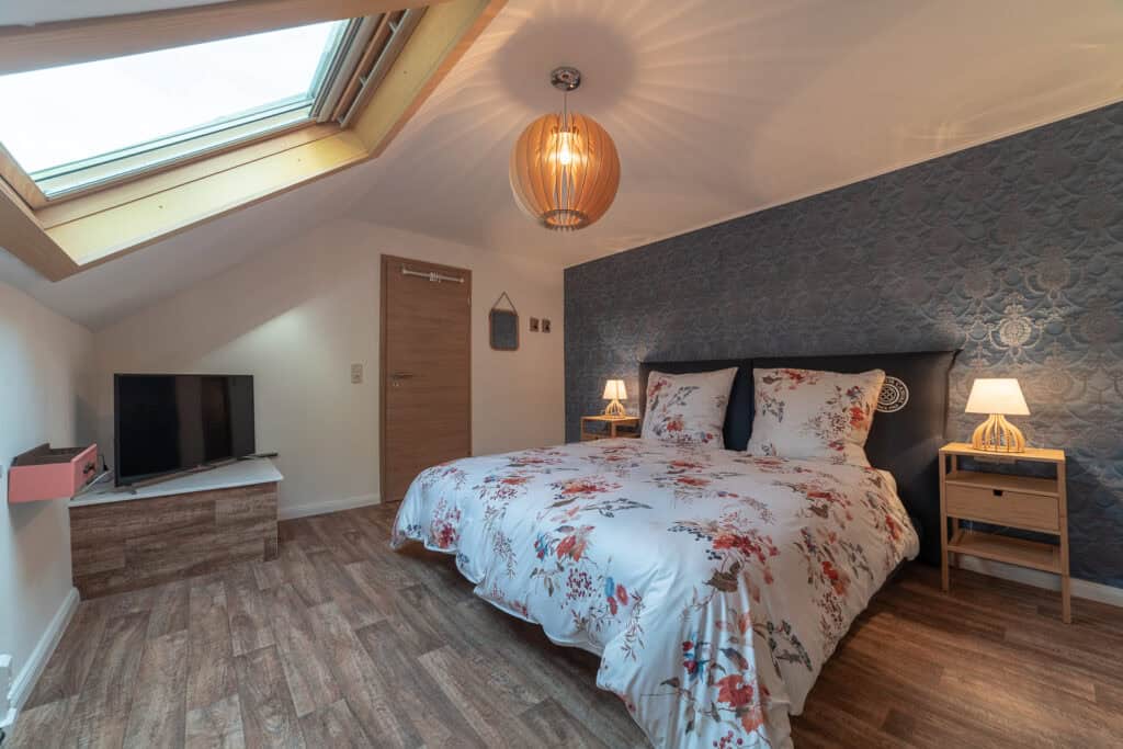 Ein gemütliches Schlafzimmer in diesem Ferienhaus Eifel verfügt über ein Bett mit Blumenmuster, zwei Nachttische mit Lampen, einen Fernseher auf einem Holzständer, Holzboden, eine blaue Akzentwand und ein großes Dachfenster, das den Raum mit natürlichem Licht füllt.