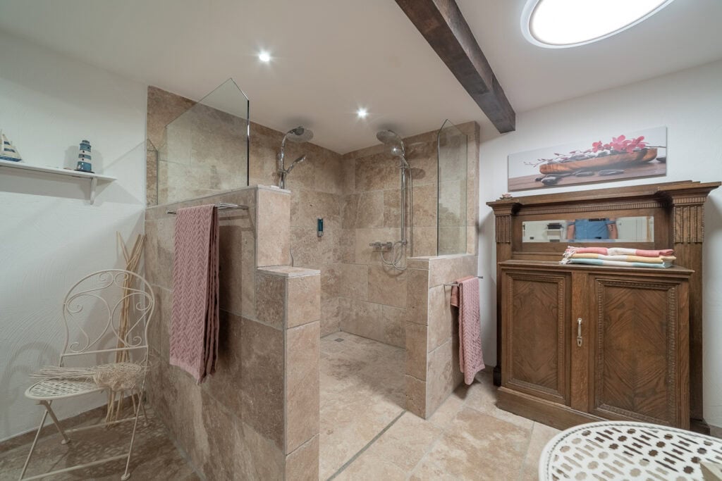Geräumiges Badezimmer in diesem Ferienhaus in Monschau mit einer begehbaren Doppeldusche, hellbraunen Steinfliesen, Glastrennwänden, einem Holzschrank mit Handtüchern, einem weißen Metallstuhl, Regaldekor, Deckenleuchten und einem charmanten Holzbalken – perfekt für Ihren Familienurlaub in der Eifel.
