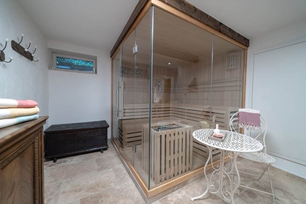 Eine moderne Sauna mit Glaswänden befindet sich in einem hellen Raum im Ferienhaus Eifel, neben einem kleinen weißen Metalltisch mit zwei Stühlen. Gefaltete Handtücher und eine Holztruhe stehen an der Wand, und der geflieste Boden rundet diesen entspannenden Raum ab.