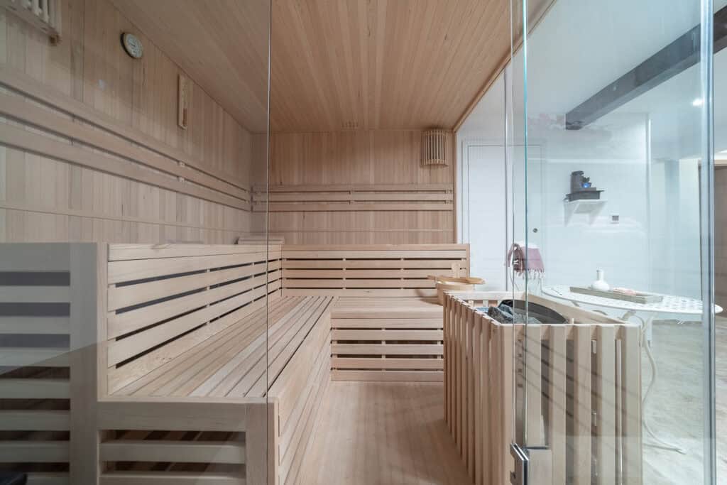 Eine moderne Holzsauna mit Sitzbänken und Glastür, angrenzend an einen hellen Raum mit Tisch und Stühlen – perfekt zum Entspannen während Ihres Eifelwochenendes oder eines Familienaufenthalts im Ferienhaus Eifel.