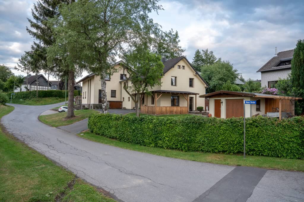 Eine ruhige Vorstadtstraße führt an einem beigefarbenen Ferienhaus Eifel mit Holzveranda vorbei, umgeben von grünen Büschen und Bäumen. In der Nähe steht ein kleiner Schuppen, ideal für Familien, die die Eifel mit Kindern erkunden, während im Hintergrund weitere Häuser zu sehen sind.
