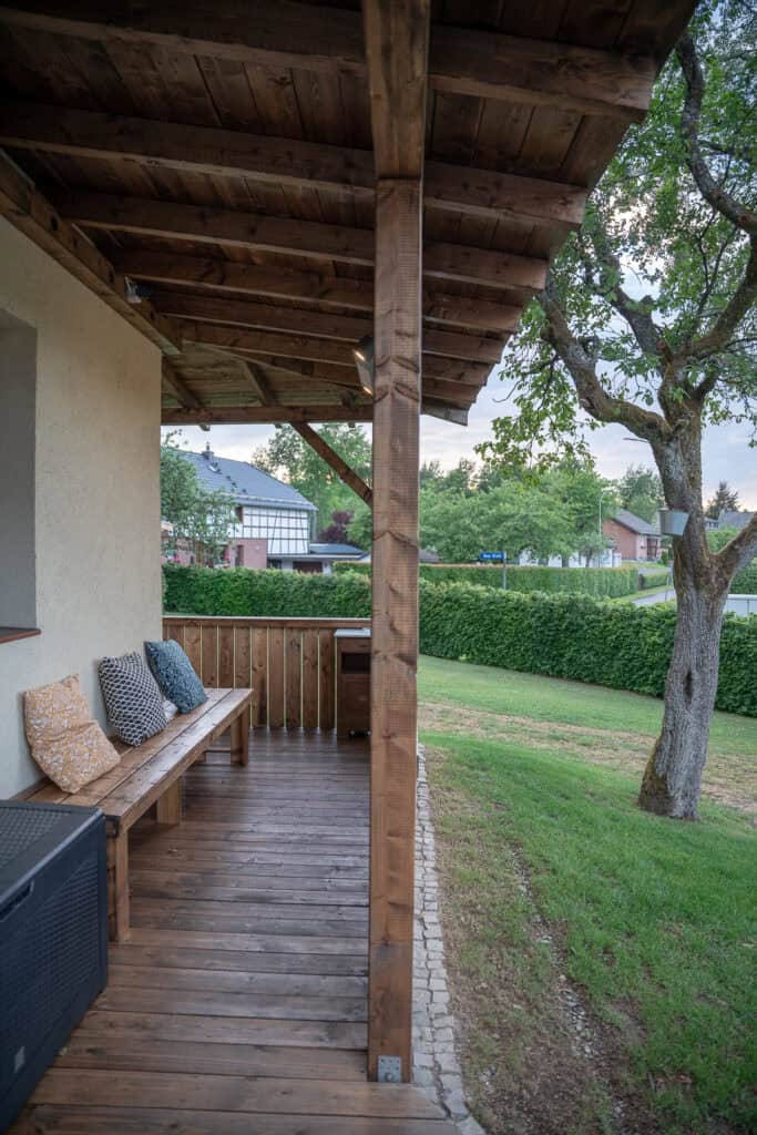 Holzveranda mit Bank und Kissen unter einem überdachten Dach, neben einem Rasenhof mit einem Baum; die perfekte Kulisse zum Entspannen in Ihrem Ferienhaus Eifel, mit Häusern und grünen Hecken im Hintergrund an einem bewölkten Tag.