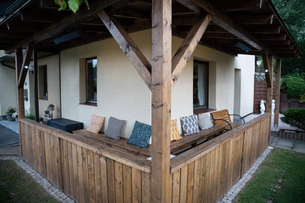 Eine überdachte Holzveranda, die an ein beiges Ferienhaus in der Eifel angebaut ist, verfügt über eine eingebaute Bank mit bunt gemusterten Kissen, einen kleinen Tisch und grünes Gras, das den Bereich umgibt – perfekt für ein Eifelwochenende.