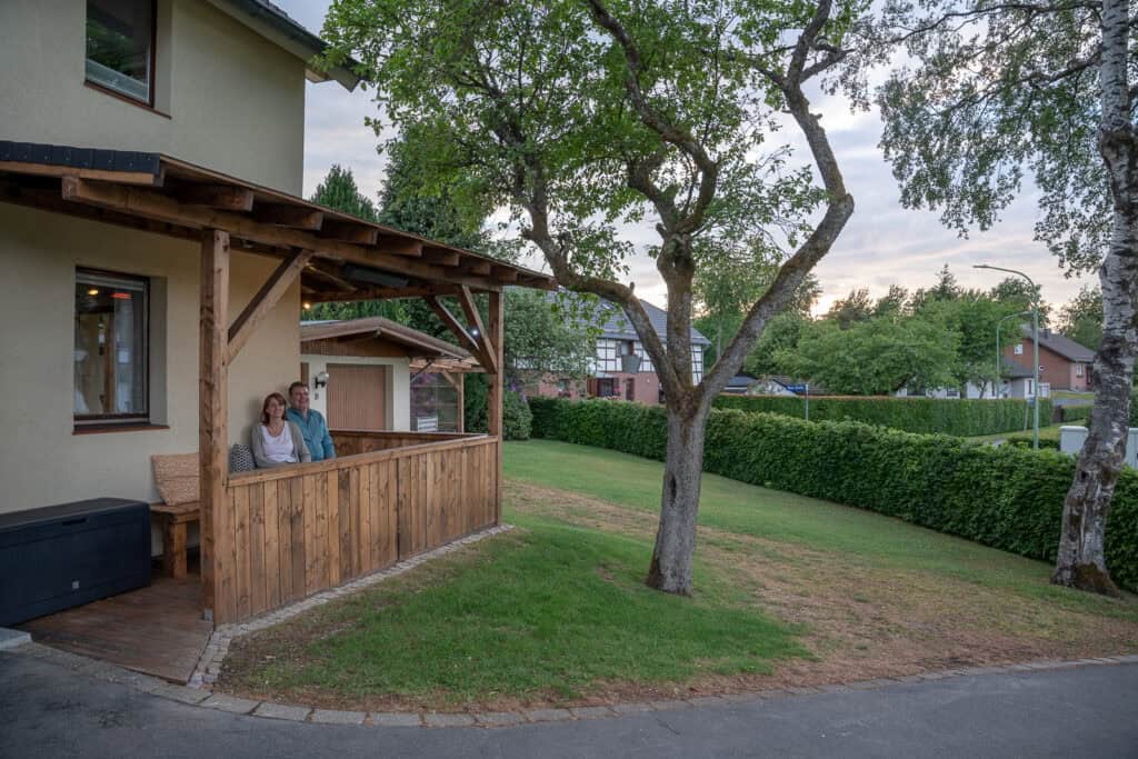 Ein Paar sitzt bei Sonnenuntergang zusammen auf der Holzveranda ihres Ferienhauses in Monschau, umgeben von grünem Gras, Bäumen und einer ruhigen Wohngegend – perfekt für ein friedliches Eifelwochenende.