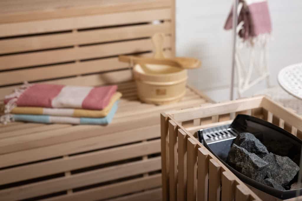 Eine gemütliche Holzsauna in Ihrem Ferienhaus in Monschau mit einer Bank mit gefalteten Handtüchern, einem Holzeimer mit Schöpfkelle und einem Ofen mit Saunasteinen – ein perfekter Rückzugsort nach der Erkundung der Eifel mit Kindern.