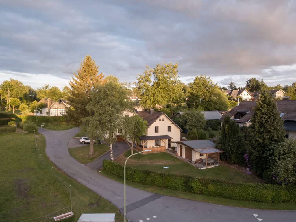 Ein ruhiges Vorortviertel mit Einfamilienhäusern, perfekt für einen Familienurlaub in der Eifel, umgeben von Bäumen und Grün, durchzogen von kurvigen Straßen unter einem bewölkten Himmel in der Abenddämmerung.