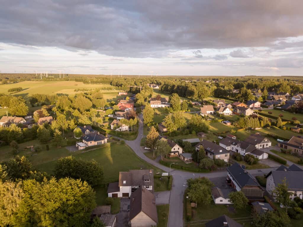 Luftaufnahme eines kleinen Dorfes mit Häusern, die zwischen grünen Bäumen und Wiesen verstreut liegen, umgeben von Feldern und Wäldern unter einem teilweise bewölkten Himmel während des Sonnenuntergangs – ideal für einen erholsamen Kurzurlaub im Ferienhaus Sauna Eifel oder ein unvergessliches Eifel-Wochenende.