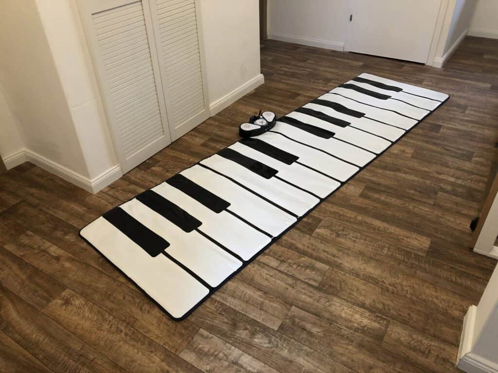 Auf dem Holzboden im Flur eines charmanten Ferienhauses in Monschau liegt eine große Spielmatte mit Klaviertasten. Am oberen Ende der Matte steht ein Paar kleine Schuhe.