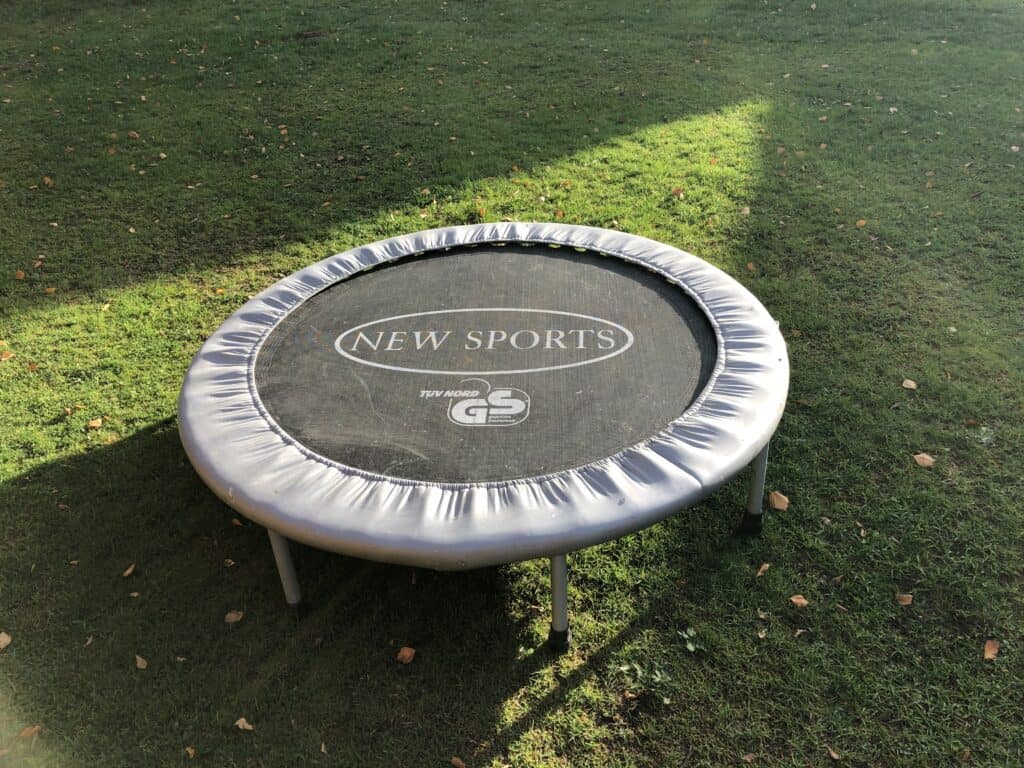 Ein rundes Outdoor-Trampolin mit den Logos „NEW SPORTS“ und „GS“ steht auf grünem Gras und wirft im Sonnenlicht Schatten – perfekt für den Spaß im Familienurlaub in der Eifel, wobei verstreute Blätter die gemütliche Atmosphäre des Ferienhauses Eifel noch verstärken.