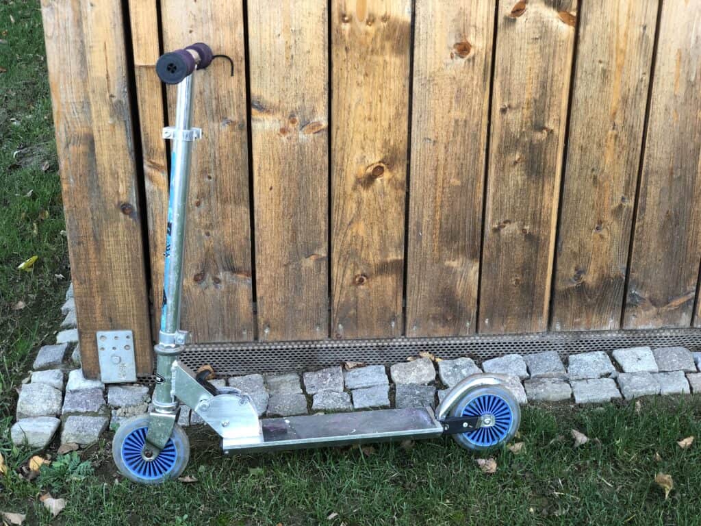 Ein silberner Tretroller mit blauen Rädern steht auf einem Kopfsteinpflasterweg neben einer Holzmauer – perfekt, um bei einem Familienurlaub in der Eifel oder einem Aufenthalt in einem Ferienhaus in Monschau die grüne Umgebung und die gefallenen Blätter zu erkunden.
