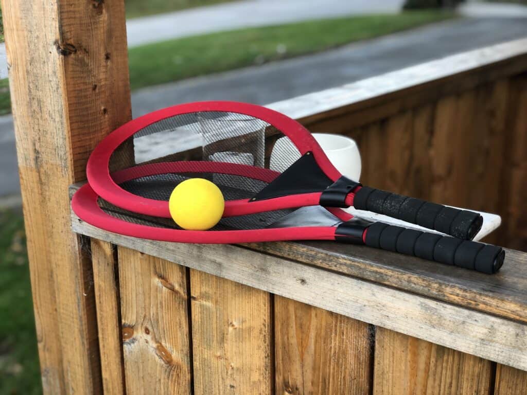 Zwei rot-schwarze Schläger mit schwarzen Griffen und einem gelben Ball liegen auf einem Holzgeländer neben einem weißen Becher, im Freien mit Gras und einer Straße im Hintergrund – perfekt für entspannte Momente im Ferienhaus Monschau.