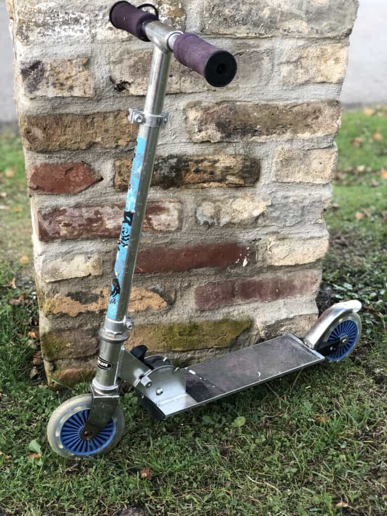 Ein silberner Tretroller mit blauen Rädern und abgenutzten lila Griffen lehnt an einer Steinmauer im Gras – perfekt für ein Eifelwochenende oder zum Erkunden der Umgebung des Ferienhauses Monschau mit Kindern. Der Roller weist Rost- und Verwitterungsspuren auf.