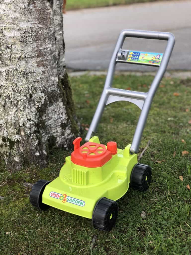 Ein leuchtend grüner und roter Spielzeug-Rasenmäher mit schwarzen Rädern steht im Gras neben einem Baum; er erinnert an den Garten eines Monschauer Ferienhauses, im unscharfen Hintergrund ist eine Straße zu sehen.