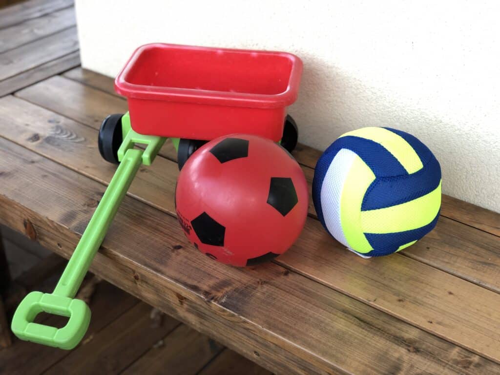 Auf einer Holzbank steht ein kleiner roter Wagen mit grünem Griff neben einem roten Fußball und einem blau-gelben Volleyball – perfekt für den Familienspaß im Ferienhaus Eifel, insbesondere wenn man die Eifel mit Kindern erkundet.