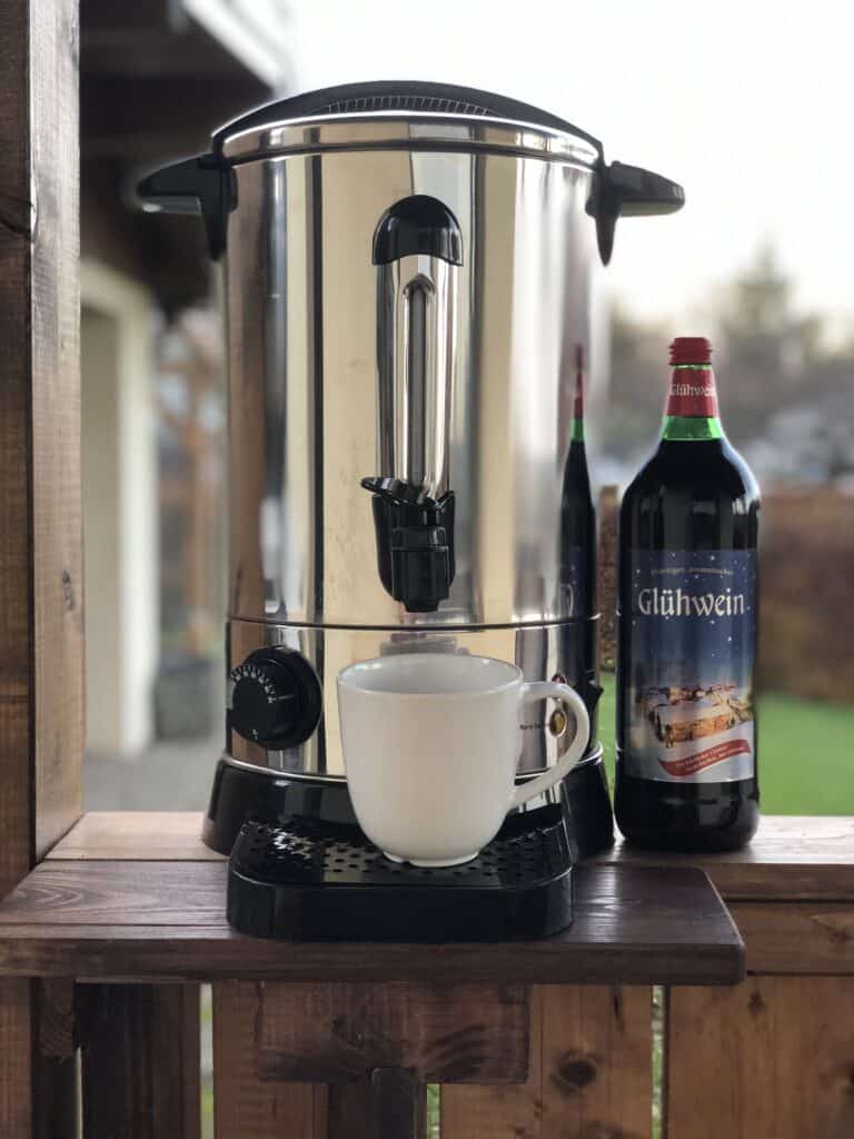 Eine große silberne Getränkekanne mit Ausguss steht auf einer Holzfläche neben einer Flasche mit der Aufschrift „Glühwein“. Perfekt für gemütliche Abende in Ihrem Ferienhaus in Monschau: Unter dem Ausguss steht ein weißer Becher, der nur darauf wartet, gefüllt zu werden, während draußen Grünpflanzen wachsen.