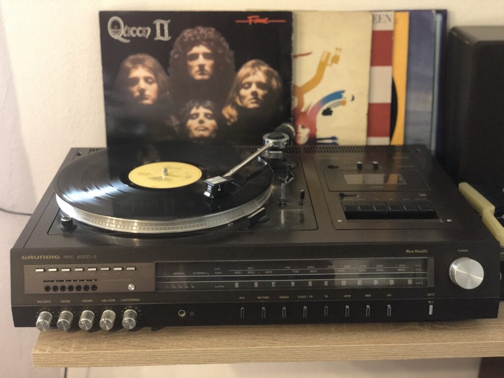 Eine Vintage-Stereoanlage von Grundig mit einer sich auf dem Plattenteller drehenden Vinyl-Schallplatte sorgt für Retro-Vibes – das perfekte Ambiente für ein entspanntes Eifelwochenende oder einen gemütlichen Abend in Ihrem Ferienhaus in Monschau, denn klassische Albumcover wie „Queen II“ verleihen zusätzlichen Charme.