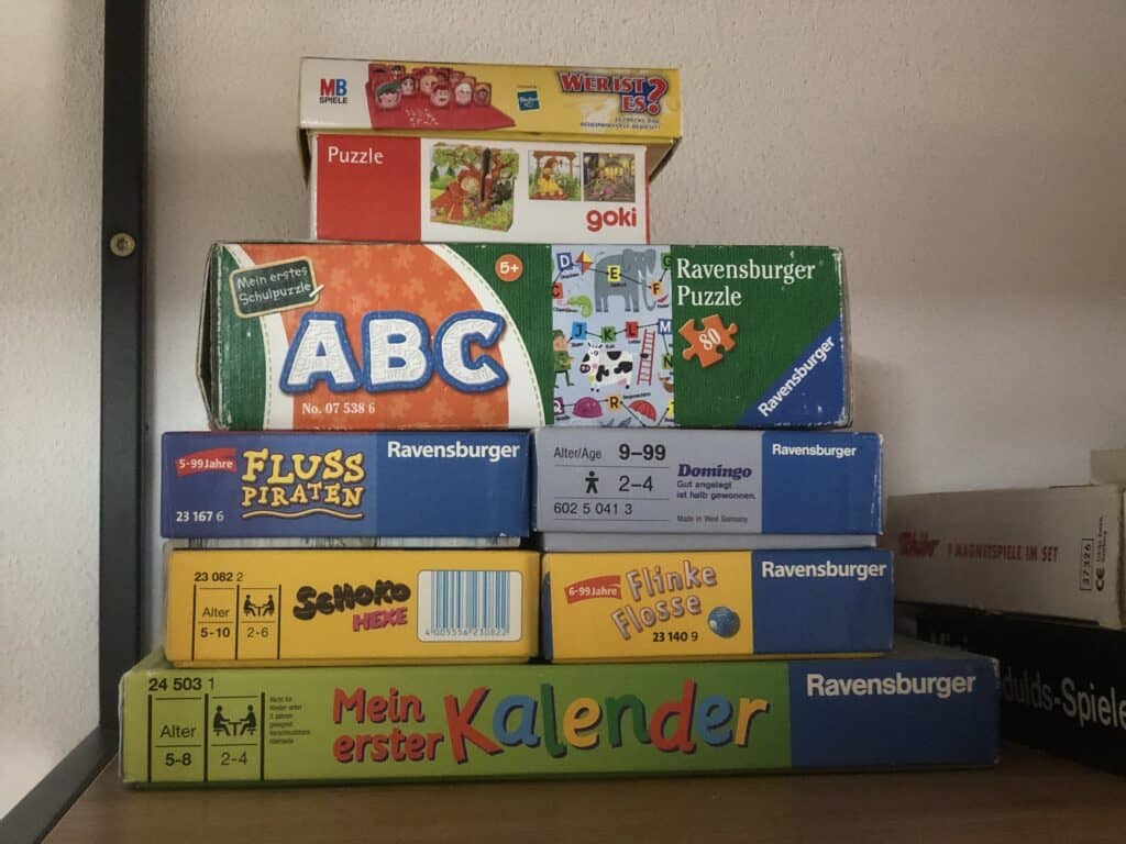 Ein Stapel bunter Ravensburger Puzzle- und Brettspielboxen steht auf einem Regal an der Wand – perfekt für ein Eifelwochenende oder gemütliche Abende in Ihrem Ferienhaus in Monschau. Die Boxen zeigen verschiedene Designs, Titel und Altersempfehlungen auf Deutsch.