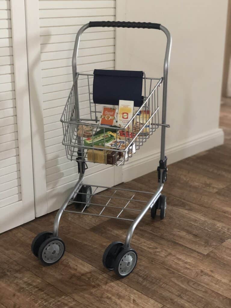 Ein kleiner Spielzeug-Einkaufswagen aus Metall mit vier Rädern, gefüllt mit Miniatur-Einkaufskartons, steht auf einem Holzboden neben einer weißen Schranktür – die perfekte Inspiration für die Planung Ihres nächsten Familienurlaubs in der Eifel.