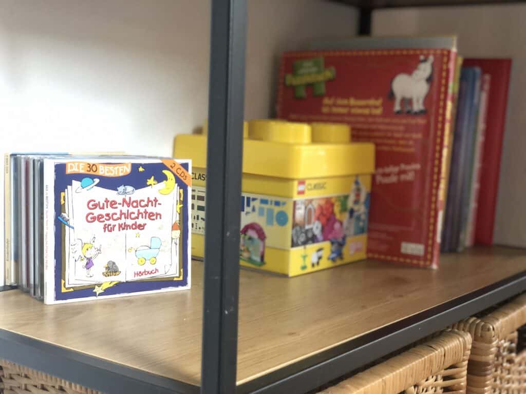 Ein hölzernes Bücherregal mit schwarzem Metallrahmen präsentiert Kinderbücher, LEGO-Aufbewahrungsboxen und die CD „Gute-Nacht-Geschichten für Kinder“ – ein gemütlicher Ort in einem Ferienhaus Eifel, perfekt für entspannte Familienmomente nach der Erkundung der Eifel mit Kindern.