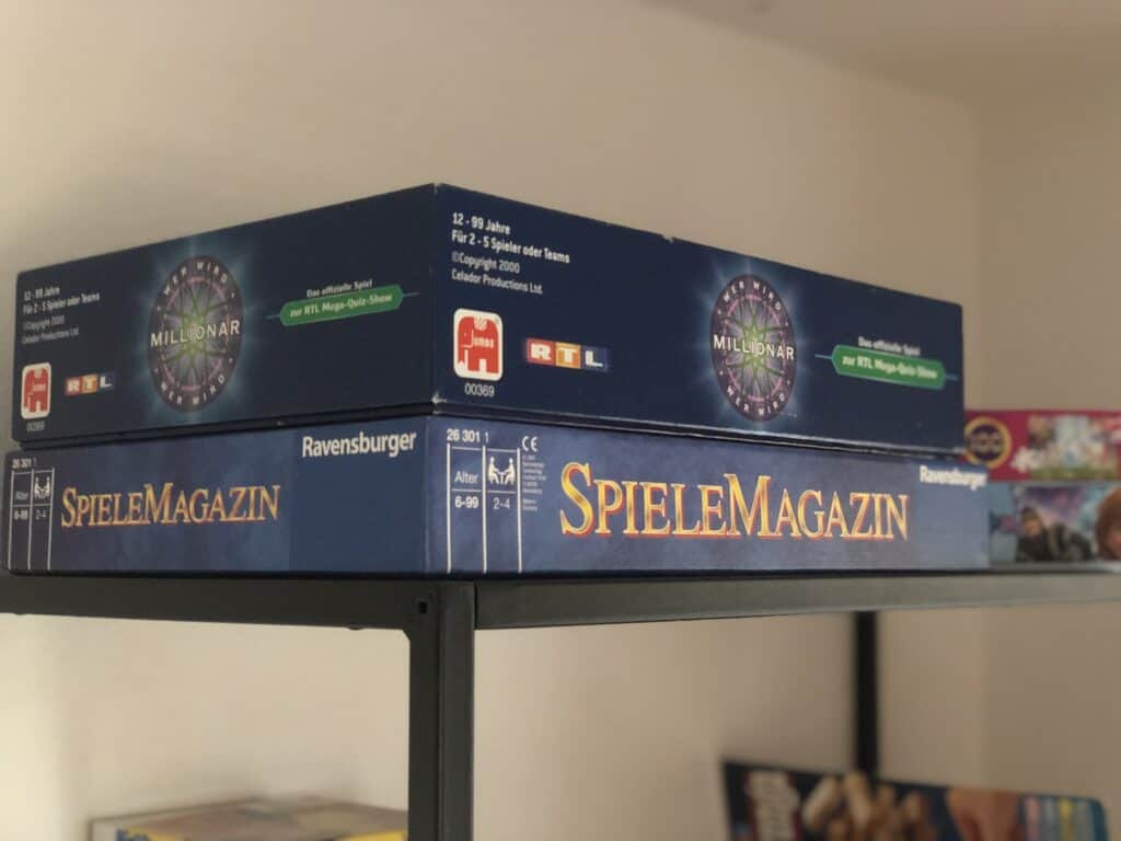 Zwei Brettspiele stapeln sich auf einem schwarzen Regal – ideal für den Familienurlaub in der Eifel. Die oberste Box trägt das Spiel „Wer wird Millionär?“ und ruht auf dem „SpieleMagazin“ von Ravensburger. Die beiden blauen Boxen sind mit weiß-gelbem Text versehen – perfekte Unterhaltung im Ferienhaus in der Eifel.