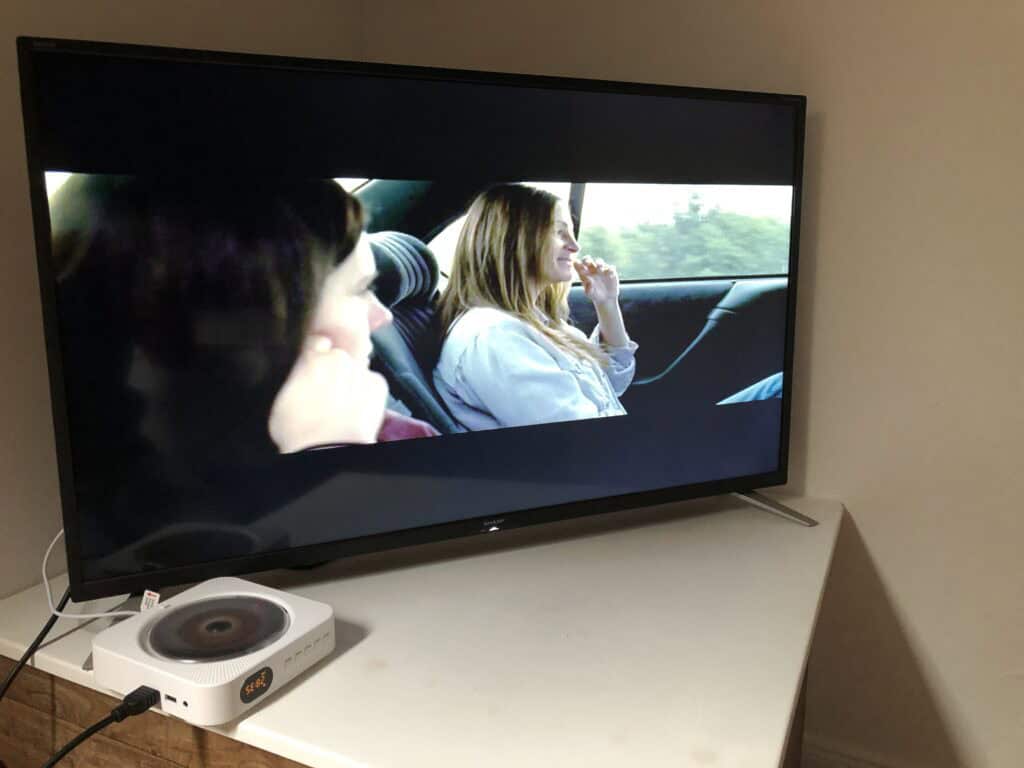 Ein Fernseher auf einem weißen Ecktisch zeigt zwei Personen auf dem Rücksitz eines Autos – perfekt für entspannte Abende während Ihres Familienurlaubs in der Eifel. Daneben steht ein kleines weißes Gerät mit einer rotierenden CD, alles vor einer schlichten, hellen Wand.