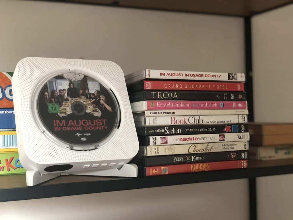Ein Regal mit einem weißen CD-Player, auf dem die CD „Im August in Osage County“ läuft. Daneben ein Stapel DVDs, perfekt für ein gemütliches Eifel-Wochenende oder entspannte Abende in Ihrem Ferienhaus Sauna Eifel.