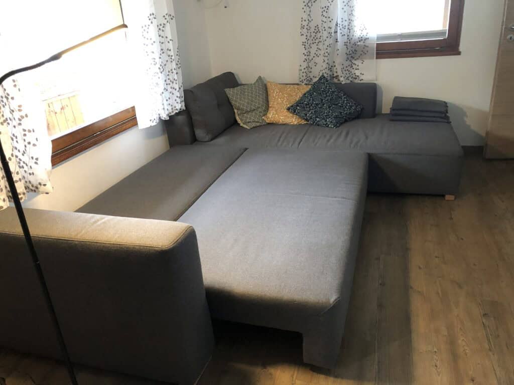 Ein graues L-förmiges Sofa mit gemusterten Kissen und gefalteten Decken steht in einem hellen Raum mit Holzböden, weißen Wänden und zwei Fenstern – perfekt zum Entspannen nach einem Familienurlaub in der Eifel oder zum Erkunden der Eifel mit Kindern.
