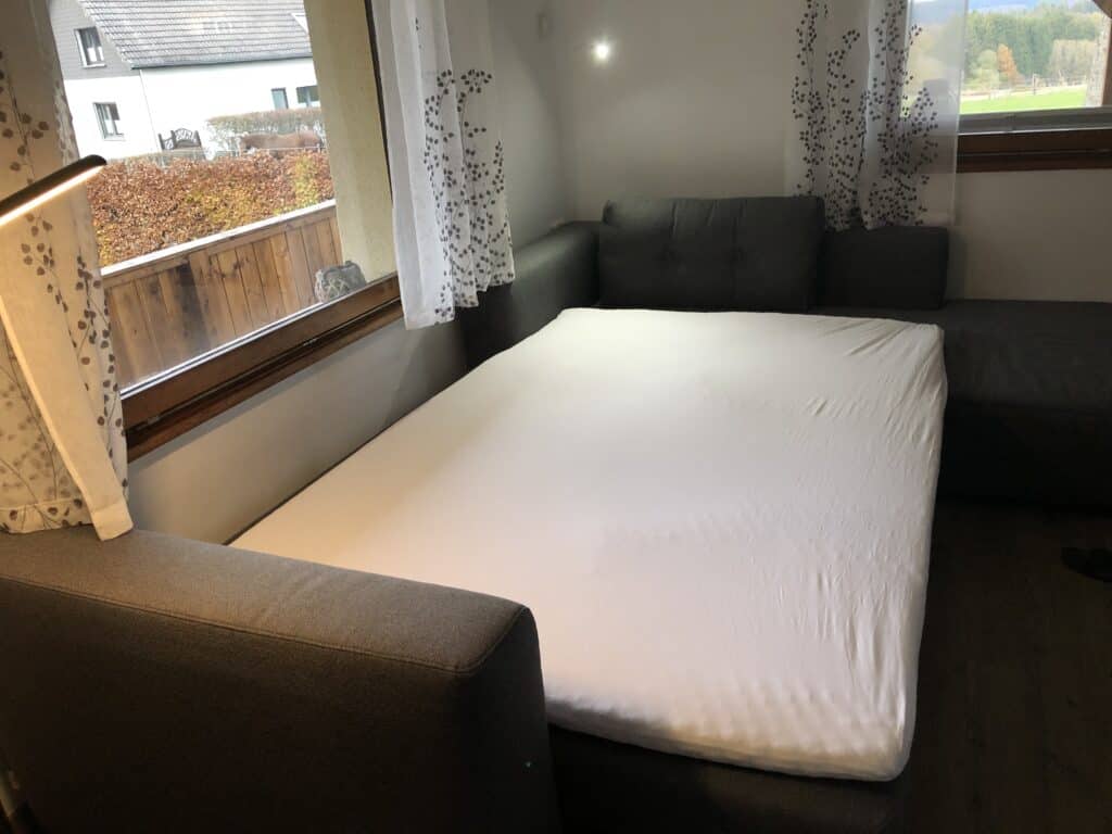 Ein graues Schlafsofa lässt sich zu einem großen Bett ausklappen, in der Nähe von Fenstern mit transparenten weißen Vorhängen. Das Zimmer ist perfekt für einen Familienurlaub in der Eifel, verfügt über Holzböden und bietet einen Blick auf Häuser und Grünflächen draußen.
