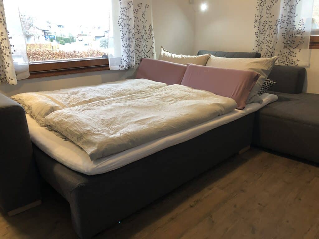Ein graues Schlafsofa mit weißer Bettdecke und rosa-weißen Kissen steht in einem gemütlichen Zimmer mit Holzboden – perfekt für ein Eifelwochenende oder entspannte Abende nach der Erkundung des Ferienhauses Eifel. Ein Fenster mit transparenten Vorhängen und ein Ecksofa runden das Ambiente ab.