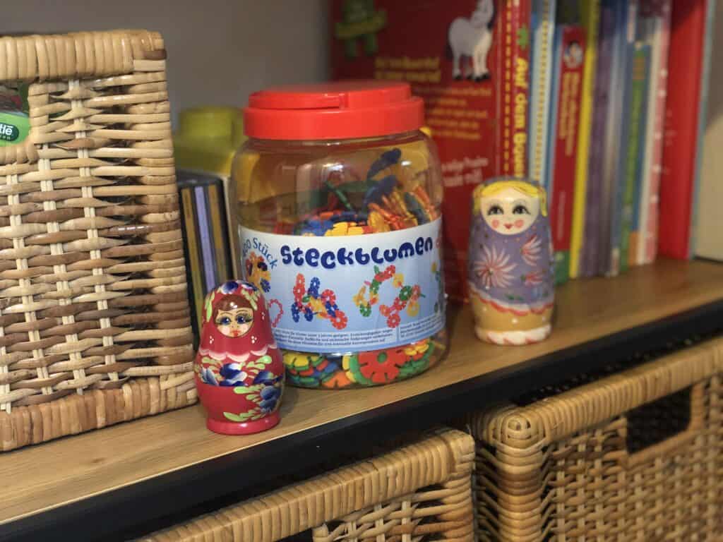 Auf einem Regal in diesem Ferienhaus in Monschau befinden sich geflochtene Körbe, ein mit bunten Stücken gefülltes „Steckblumen“-Glas, russische Matrjoschkas und Kinderbücher – perfekte Details für einen Familienurlaub in der Eifel.