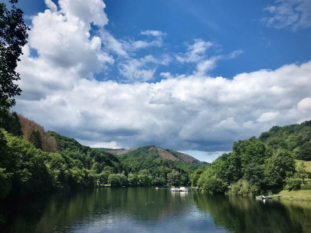 Ein ruhiger Fluss, gesäumt von üppigen grünen Bäumen mit sanften Hügeln im Hintergrund unter einem teilweise bewölkten blauen Himmel; perfekt für ein Eifel-Wochenende oder einen erholsamen Urlaub in einem Ferienhaus Sauna Eifel, mit kleinen Booten, die sanft auf dem Wasser treiben.