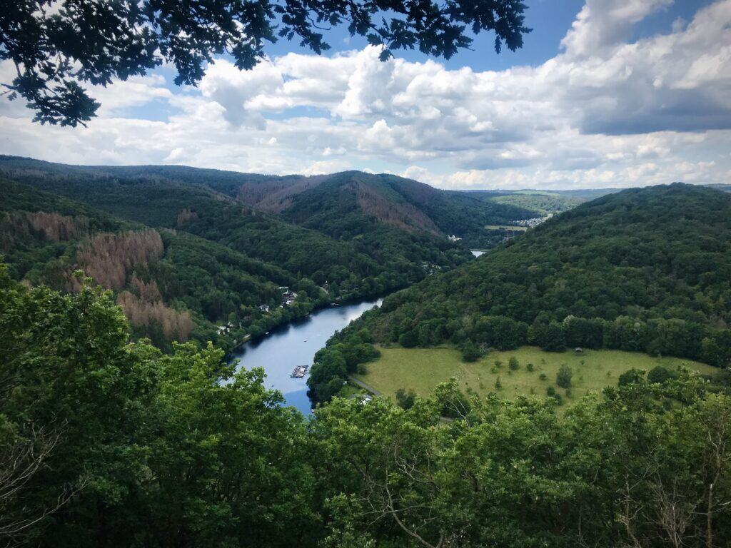 Ein Fluss schlängelt sich unter einem teilweise bewölkten Himmel durch üppig grüne Hügel und Wälder in der Region Eifel mit Kindern. Auf dem Wasser sind sonnenbeschienene Grasflächen und kleine Boote zu sehen.