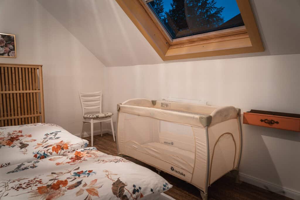 Ein gemütliches Kinderzimmer in einem Ferienhaus in Monschau verfügt über ein Babybett, geblümte Bettwäsche auf einem Bett, einen weißen Stuhl, einen Holzschrank, ein kleines Wandregal und ein Dachfenster, durch das man den Abendhimmel und die Äste draußen sehen kann.