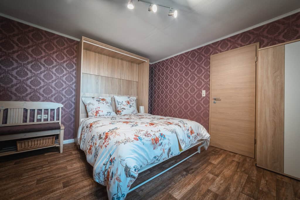 Ein gemütliches Schlafzimmer mit gemusterter Tapete, einem Doppelbett mit geblümter Bettwäsche, Holzböden und einer gepolsterten Bank – perfekt für einen Familienurlaub in der Eifel oder zum Entspannen nach einer Erkundungstour durch die Eifel mit Kindern unter sanften Deckenstrahlern.