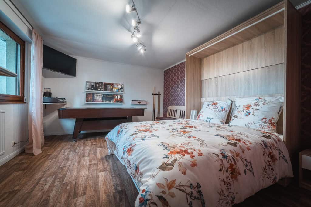 Ein gemütliches Schlafzimmer mit Holzboden, geblümter Bettdecke auf einem Doppelbett, Wandfernseher, Tischfußball und Schallplattenregal – perfekt für einen Familienurlaub in der Eifel. Sanfte Beleuchtung und ein großes Fenster verleihen diesem einladenden Ferienhaus in der Eifel Wärme.