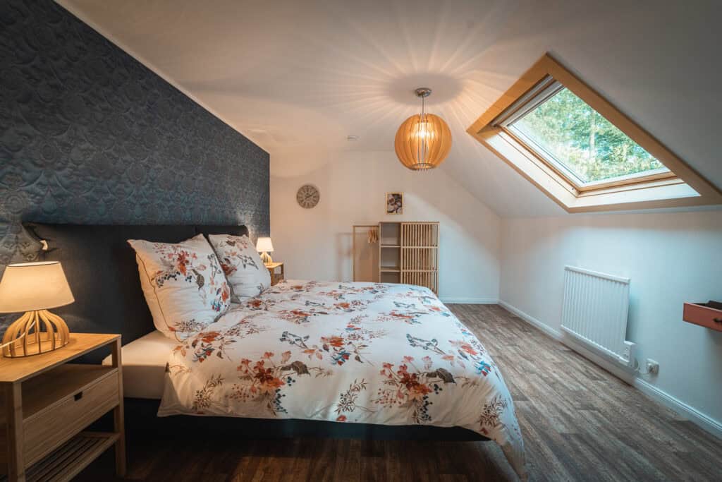Ein gemütliches Dachgeschoss-Schlafzimmer in einem Monschauer Ferienhaus mit geblümtem Bett, hölzernen Nachttischen, warmer Beleuchtung, Dachfenster und Holzboden. Das Zimmer ist modern eingerichtet und bietet eine ruhige, luftige Atmosphäre.