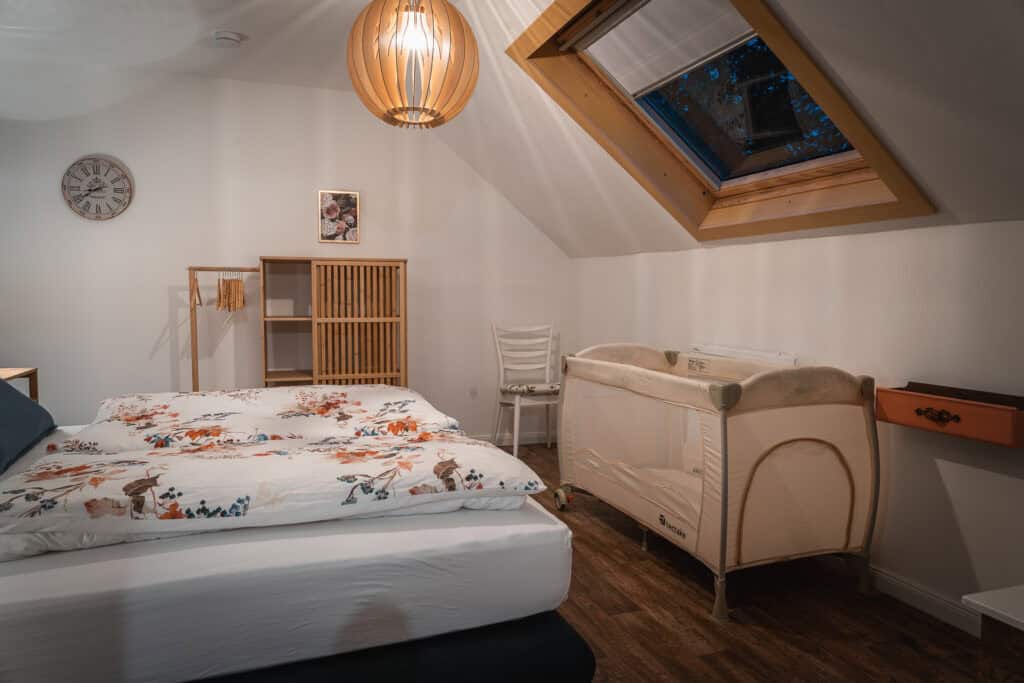 Ein gemütliches Schlafzimmer mit einem Doppelbett, geblümter Bettwäsche, einem Holzbettchen, einem Dachfenster und einfachen Holzmöbeln auf einem dunklen Holzboden – perfekt für Familien, die das Ferienhaus Eifel oder einen Kurzurlaub in der Eifel mit Kindern genießen.