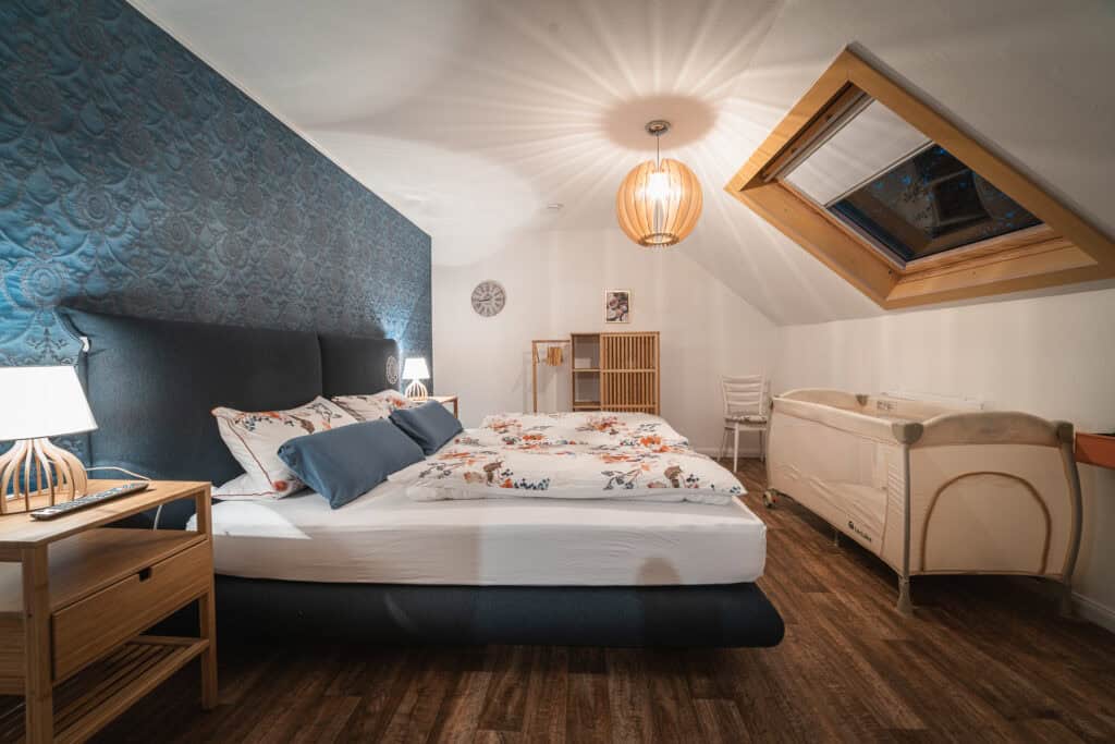 Ein gemütliches Schlafzimmer in einem Ferienhaus in der Eifel mit Doppelbett, geblümter Bettwäsche, blauer Akzentwand, Nachttischen mit Lampen, Holzboden, Hängelampe, Dachfenster, Kinderbett und minimalistischen Holzmöbeln – ideal für einen Familienurlaub in der Eifel.