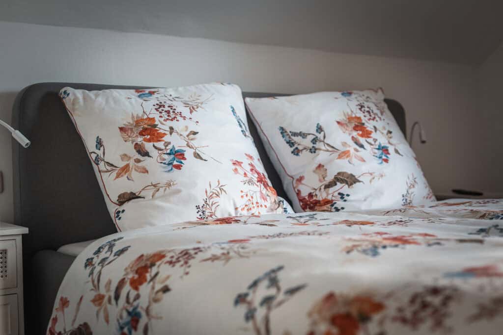 Ein ordentlich gemachtes Bett mit grauem Kopfteil und weißer Bettwäsche mit farbenfrohen Blumenmustern. Zwei große Kissen passen perfekt zur Bettdecke, und an beiden Seiten sind Leselampen angebracht – perfekt zum Entspannen in Ihrem Monschau Ferienhaus nach einer Entdeckungsreise durch die Eifel mit Kindern.